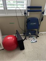 Photo shows a red yoga ball, blue chair gym with padded seats and resistance bands labeled RAD BISHOP, and black foam wedge against a wall with window.