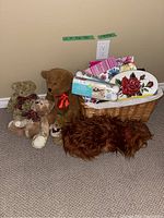 Four stuffed teddy bears and two stuffed dogs arranged around a wicker basket filled with floral and striped gift bags, two Easter egg decorating kits with white eggs, and a decorative tin with rose design.