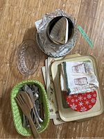Overview of kitchen items including cutlery basket, glass serving dishes, metal trays, linens, placemats and a small red cookie tin.