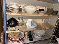 Shelf with multiple stacked white ceramic plates, bowls, glass dishes, black Crock-Pot, and small square dishes