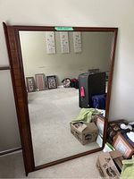 Large rectangular mirror with clear glass and narrow wooden frame leaning against a wall in a room with various other items visible in reflection.