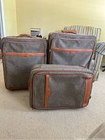 Three Hartmann tweed and leather rolling suitcases arranged standing and front-facing showing size variation.