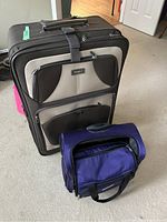 Medium shot showing large black and gray soft-sided luggage and smaller dark purple carry-on bag seated on a carpeted floor.
