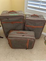 Three Hartmann tweed and leather rolling suitcases arranged on carpeted floor, shows full set of small, medium, and large sizes