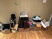 Wide view of a large assortment of office supplies stacked in boxes and organizers on a hardwood floor against a plain wall. Visible items include binders, paper organizers, and various office supplies.