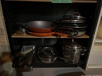 Cookware set on shelving unit including orange handle frying pans, stainless steel pots with lids, tongs, and skewers
