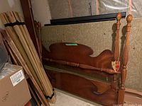 Disassembled Gibbard queen four poster bed frame parts including headboard, footboard, four posts with spindle design, and bundled wooden slats leaning against wall in basement.