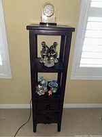 Wooden display shelf with decorative items on shelves including silver-tone figurines, cat figurines, Wedgwood ceramics, and a fossil.
