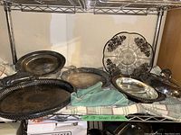 Multiple silver plated trays including oval and round with patterns, a covered dish with glass insert, a glass plate with floral design on a shelf