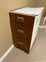 Front view of small three-drawer filing cabinet showing wood laminate drawers with metal handles and grid paper on top.
