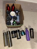 Top view of open cardboard box containing various cups and water bottles, plus several thermal mugs and containers arranged on floor outside box.