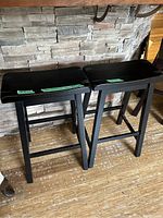 Two black wooden bar stools placed side by side on wooden floor against a stone wall background, showing tops and legs with visible wear.