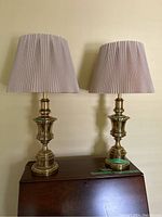 Two brass Stiffle table lamps with pleated beige shades, placed side by side on a wooden surface against a wall.