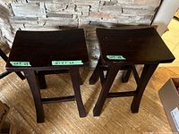 Two brown wooden barstools side by side on a wooden floor against a stone wall, showing top and side views of seats and legs.