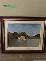 Framed coastal scene painting showing water, building, hills, and sky, framed in wood with cream and white mats.