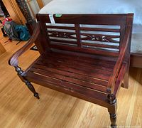 Front right angle view of dark wood bench with carved backrest and slatted seat on hardwood floor.