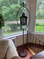 Full view of the tall black metal candle stand with circular base and oval frame, holding the lantern in front of window and couch.