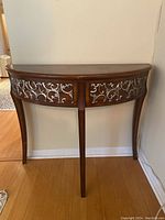 Full front view of the brown half-moon shaped console table with decorative metal scrollwork inset on the front panel and three legs.