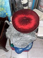 Photo showing round red velvet cushion on top of a silver/grey plush throw blanket with visible portions of furniture and storage items around.