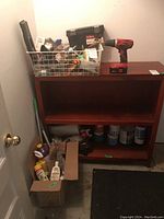 Image of wooden shelf with paints, basket of small hardware items and red and black Skil drill on top, cleaning products and a broom in boxes on floor