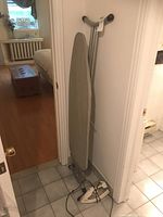 Ironing board upright against wall near door, with iron placed on floor beside it; room has tiled floor and visible radiator and furniture in adjacent room.