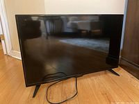 Front view of black Insignia 32 inch LED TV on hardwood floor with two stand legs and power cord visible.