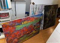 Four oil paintings leaning against a wall in a room. One painting is a colorful autumn landscape predominantly red and green, the others appear abstract.
