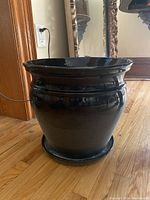 Glossy black ceramic planter with drip tray showing full height and rounded shape on hardwood floor near an electrical outlet and wooden trim.