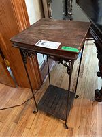Full side perspective of the wood top side table with metal base on hardwood floor with decorative metalwork on corners and thin metal legs curving at feet.