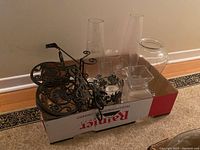 Photo of lot showing assorted glass vases including clear and floral patterned, metal decorative trivet, and metal holder in a box.