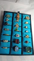 Photo showing 21 assorted vintage rings set in blue foam display, featuring multiple colorful stones and crystals in gold and silver plated bands.