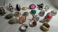 Photo showing 21 assorted rings made from non precious metals with various stones including crystals, glass, and simulated opals. Displayed on light background.