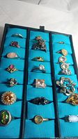 Overview of 21 rings arranged in a black display case with blue slots, showing variety of shapes, sizes, and colored stones in gold and silver toned non-precious metal rings.