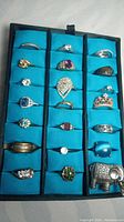 Photo shows 21 vintage used rings with various stones including crystals, glass, opals, and synthetic rubies in gold and silver tone metal settings on a blue display tray.