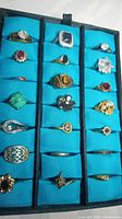 Photo showing 21 assorted non-precious rings in blue organizer tray, variety of stones including clear crystals, red synthetic stones, turquoise stones, opal-like clusters, and various metal plating.