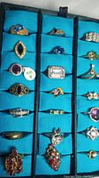 Top view of 21 assorted rings in a ring display case on turquoise lining, showing variety of styles and stones.