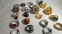 Photo showing an angled overhead view of 21 assorted vintage non-precious rings in silver and gold tones on white background. Featuring various shapes and colored stones including clear crystals, tiger’s eye-like, purple, green, and red stones, with different metal plating and finishes. Some rings have intricate band textures and stone settings, some appear glued.