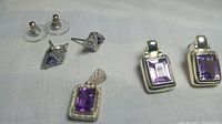 Top overview of two pairs of amethyst earrings and one amethyst pendant on a white cloth background.