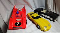 Three vintage toy cars displayed on a white background: red race car, yellow sports car, black convertible Corvette.