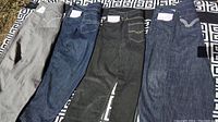 Four pairs of mens jeans laid out, showing various colors from light gray to indigo blue, with visible brand tags and typical denim styling.