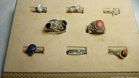 Full view of 8 sterling silver rings on beige felt display board, showing stones, shapes, and ring details.