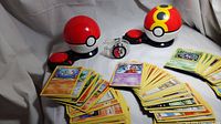 Two closed Pokemon Surprise Attack Pokeballs with piles of yellow bordered Pokemon cards fanned out in front, plus a clear band electronic Pokemon watch.