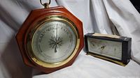 Photo showing the octagonal wooden framed barometer with brass bezel alongside the rectangular barometer with black case and brass base on white background.