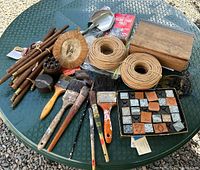 Full assortment of vintage paint brushes, bundles of rush seat repair materials, a wooden box with stamps and book, pipe cleaners, metal cow ink stamp, numerical metal stamp set, receipt holders, and leather stamps on green table