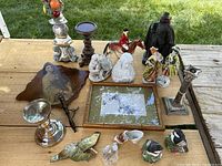 Photo showing a variety of antique decorative items on wooden surface including ceramic figurines, candlesticks, brass ornaments, and framed artwork.