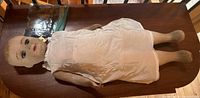 Full view of 28 inch antique cloth covered cardboard doll laying flat on table with white dress and bag of human hair placed next to head.