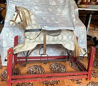 Full side view of the antique rocking horse showing the white wooden horse figure with leather ears, mane, tail, and saddle, mounted on curved metal supports attached to red wooden rockers.