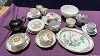 Full assortment of antique china pieces displayed on purple cloth, including black teapot, plates, cups, saucers, and platter.