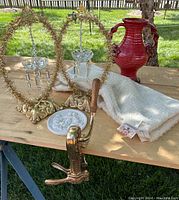 Overview of lot on table showing brass vintner clamp, red pottery vase, white plaster angel plaque, gold chandelier hangings, and pillow sham