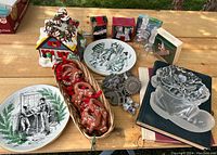 Overview of lot items including light-up Santa house decoration, basket with clay ornaments, two Charles Dickens Shenango plates, assorted pastry molds, glass Christmas shoe dish, and books.